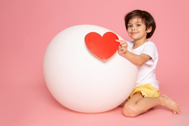 Een vooraanzicht die leuke jongen in het witte t-shirt spelen met witte bal op het roze bureau glimlacht