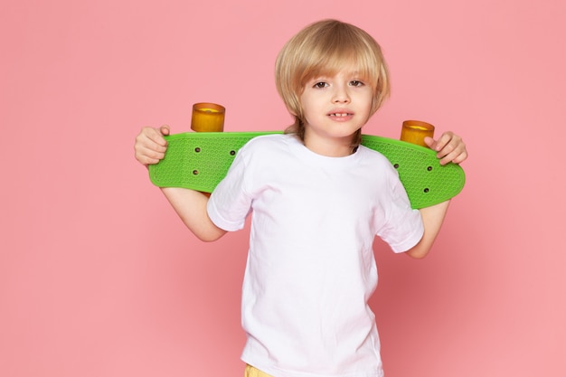 Een vooraanzicht glimlachende blondejongen die in witte t-shirt groen skateboard op de roze ruimte houdt