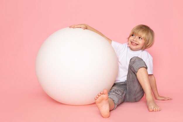 Een vooraanzicht glimlachende jongen in het witte t-shirt spelen met witte ronde bal op de roze vloer