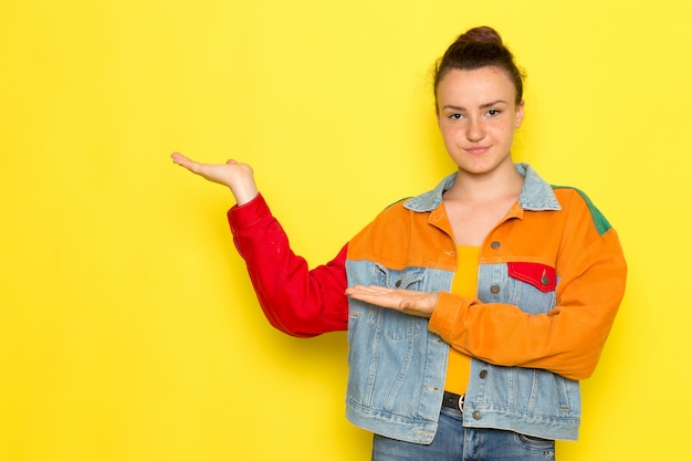 Een vooraanzicht jong wijfje in geel overhemd kleurrijk jasje en jeans die met glimlach stellen