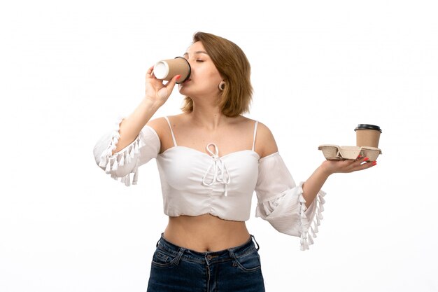 Een vooraanzicht jonge aantrekkelijke dame die in wit overhemd en jeans koffie op het wit drinkt