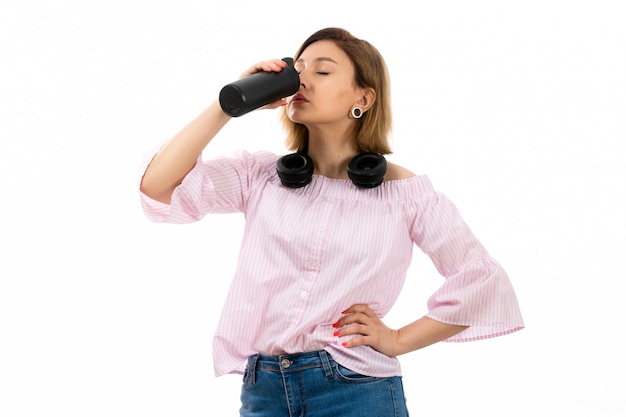 Een vooraanzicht jonge aantrekkelijke dame in roze shirt en blauwe spijkerbroek met zwarte koptelefoon drinken zwarte thermos drinken uit thermos op het wit te drinken