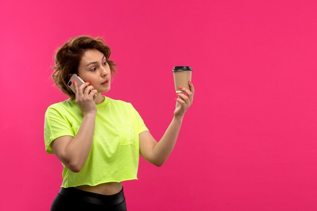 Een vooraanzicht jonge aantrekkelijke vrouw in zuur gekleurde shirt zwarte broek praten aan de telefoon met koffiekopje op de roze achtergrond jonge vrouwelijke technologieën