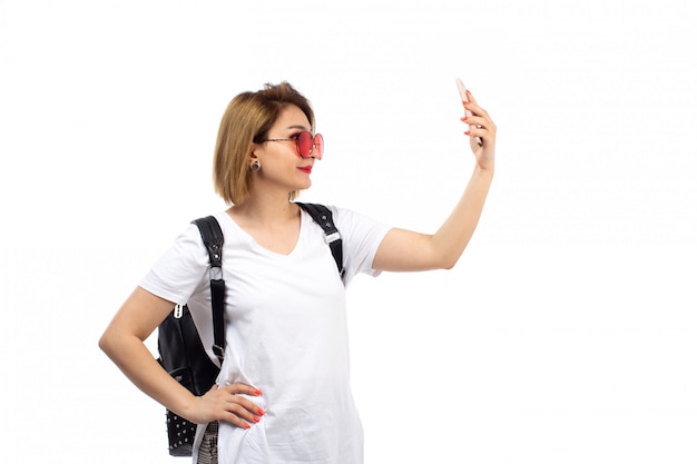 Een vooraanzicht jonge dame in witte t-shirt rode zonnebril zwarte tas glimlachend nemen selfie op het wit