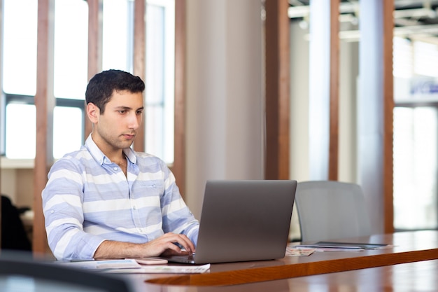 Een vooraanzicht jonge knappe mens in gestreept overhemd die binnen conferentiezaal werken die zijn zilveren laptop met behulp van tijdens de dag werkzaamhedenactiviteit