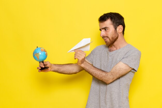 Een vooraanzicht jonge man in grijs t-shirt met kleine wereldbol en papieren vliegtuigje op de gele muur man kleurmodel