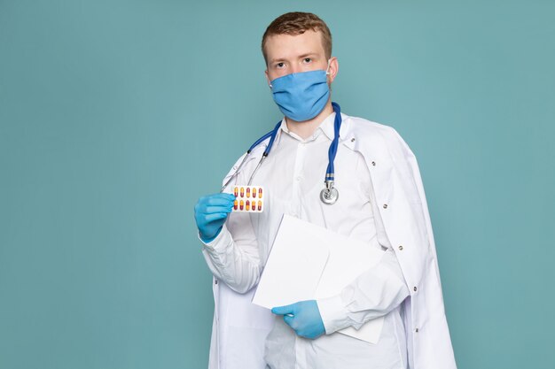 Een vooraanzicht jonge man in witte medische pak blauwe handschoenen en masker met pillen op de blauwe vloer