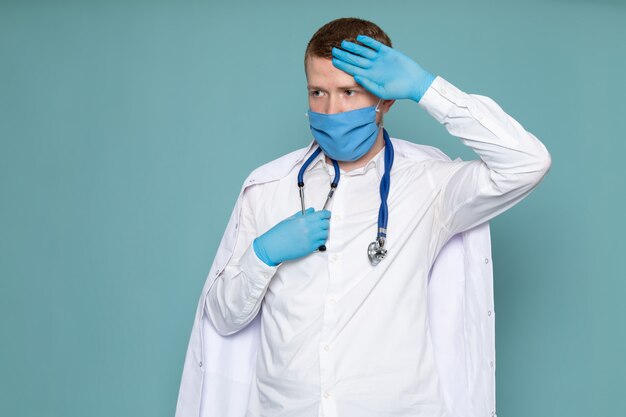 Een vooraanzicht jonge man in witte medische pak blauwe handschoenen en masker op de blauwe vloer