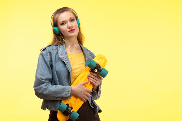 Een vooraanzicht jonge moderne vrouw in de gele overhemd zwarte broek en jeans van de de laagholding van Jean skateboard met het gekleurde oortelefoons glimlachen