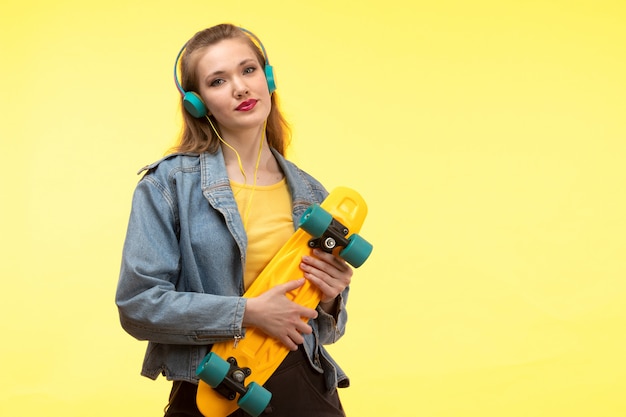 Een vooraanzicht jonge moderne vrouw in de gele overhemd zwarte broek en jeans van de de laagholding van Jean skateboard met het gekleurde oortelefoons glimlachen