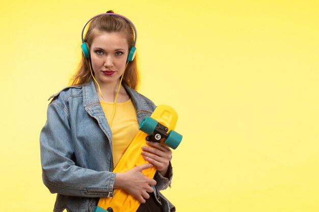 Een vooraanzicht jonge moderne vrouw in geel overhemd zwarte broek en jean jas met skateboard met gekleurde koptelefoon poseren