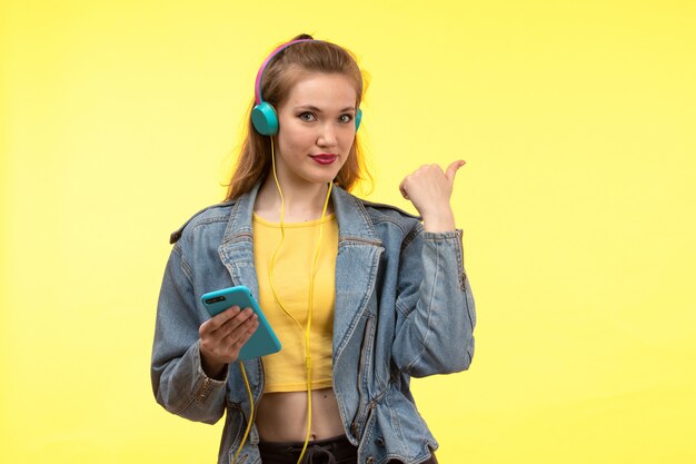 Een vooraanzicht jonge moderne vrouw in gele overhemd zwarte broek en Jean jas met gekleurde koptelefoon met behulp van telefoon poseren