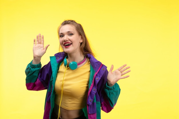 Een vooraanzicht jonge moderne vrouw in gele overhemd zwarte broek en kleurrijk jasje met gekleurde oortelefoons die gelukkige uitdrukking stellen