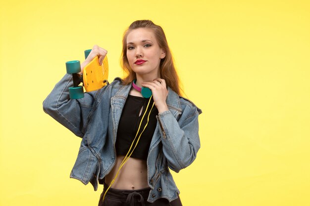 Een vooraanzicht jonge moderne vrouw in zwarte overhemd zwarte broek en jean jas met skateboard met gekleurde koptelefoon