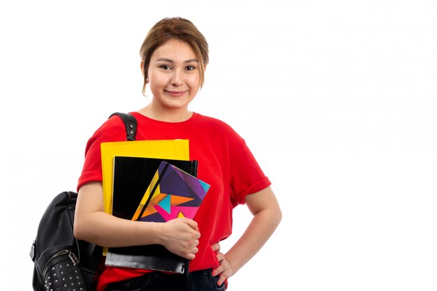 Een vooraanzicht jonge mooie dame in rode t-shirt zwarte jeans die verschillende voorbeeldenboeken en dossiers met zak op het wit glimlachen