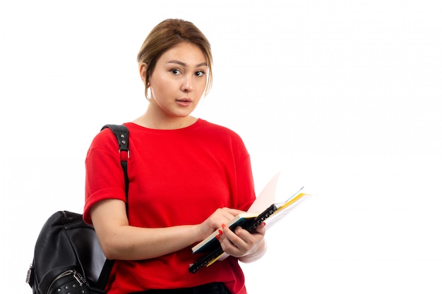 Een vooraanzicht jonge mooie dame in rode t-shirt zwarte jeans die verschillende voorbeeldenboeken en dossiers met zak op het wit houdt