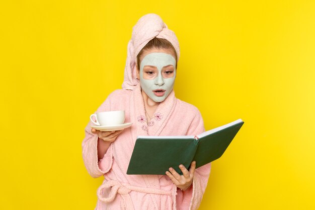 Een vooraanzicht jonge mooie vrouw in roze badjas bedrijf kopje thee leesboek