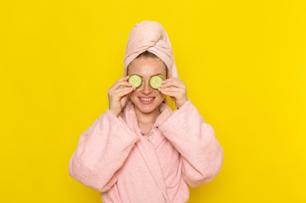 Een vooraanzicht jonge mooie vrouw in roze badjas die haar ogen bedekt met plakjes komkommer met een glimlach
