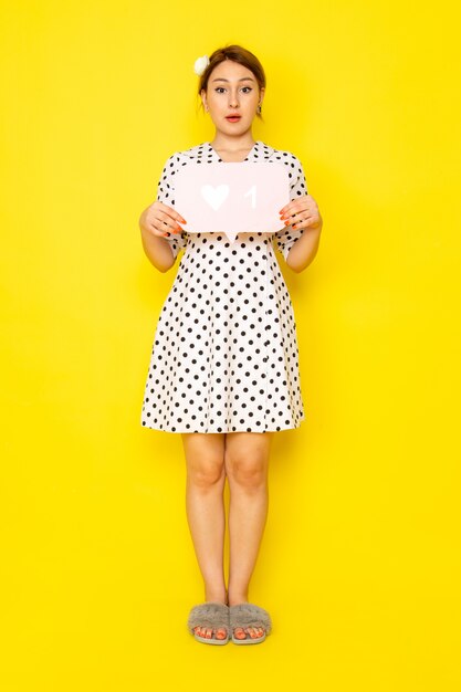 Een vooraanzicht jonge mooie vrouw in zwart-witte polka dot jurk met wit bord op geel