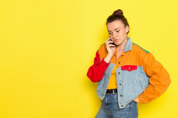 Een vooraanzicht jonge vrouw in gele overhemd kleurrijke jas en spijkerbroek praten aan de telefoon