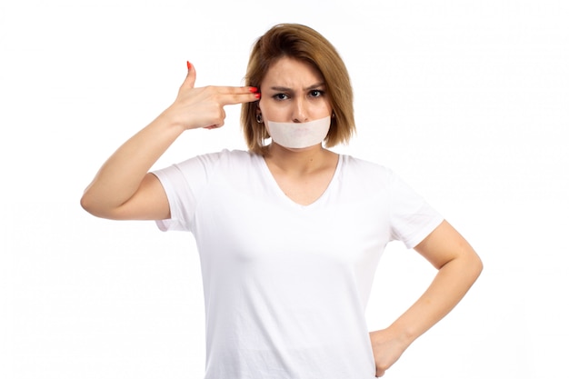 Een vooraanzicht jonge vrouw in wit t-shirt en zwarte spijkerbroek met wit verband rond haar mond met pistool op het hoofd op de witte