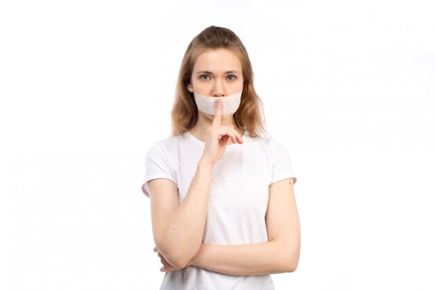 Een vooraanzicht jonge vrouw in wit t-shirt met wit verband rond haar mond met stop praten teken op de witte