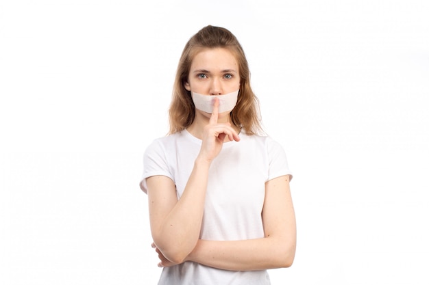 Een vooraanzicht jonge vrouw in wit t-shirt met wit verband rond haar mond met stop praten teken op de witte