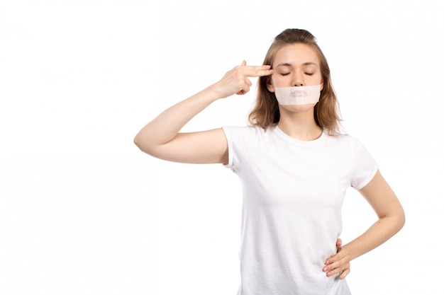 Een vooraanzicht jonge vrouw in wit t-shirt met wit verband rond haar mond wijzende vingers in haar hoofd op de witte