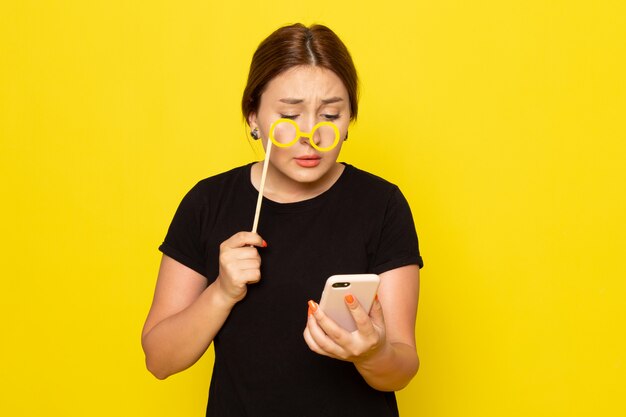 Een vooraanzicht jonge vrouw in zwarte jurk poseren met smartphone en speelgoed stok zonnebril op geel