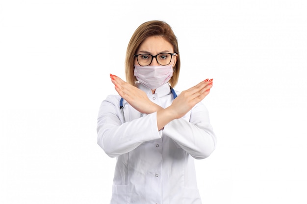 Een vooraanzicht jonge vrouwelijke arts in witte medische pak met stethoscoop dragen witte beschermend masker met verbod teken op het wit
