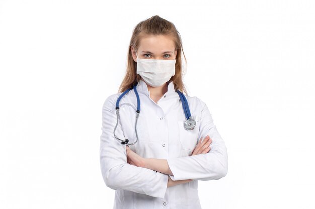 Een vooraanzicht jonge vrouwelijke arts in witte medische pak met stethoscoop dragen witte beschermend masker op de witte