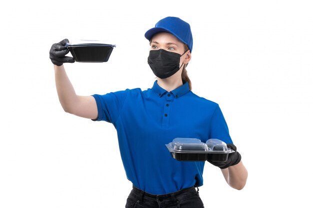 Een vooraanzicht jonge vrouwelijke koerier in blauwe uniforme zwarte handschoenen en zwart masker met voedselleveringskommen