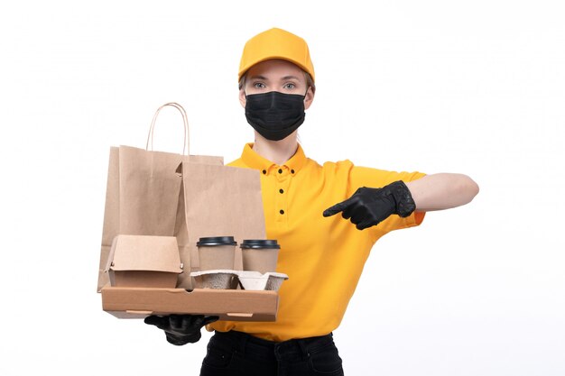 Een vooraanzicht jonge vrouwelijke koerier in gele uniforme zwarte handschoenen en zwart masker met koffiekopje voedselpakketten