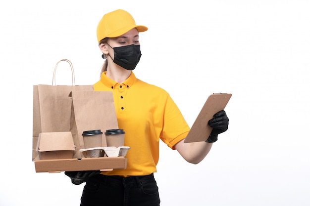 Een vooraanzicht jonge vrouwelijke koerier in gele uniforme zwarte handschoenen en zwart masker met voedselpakketten en koffiekopjes