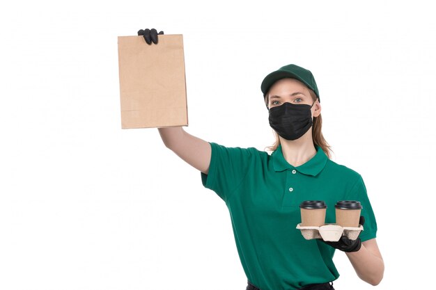 Een vooraanzicht jonge vrouwelijke koerier in groene uniforme zwarte handschoenen en zwart masker met koffiekoppen en pakket