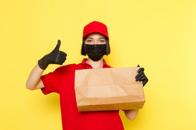 Een vooraanzicht jonge vrouwelijke koerier in rood uniform zwart handschoenen zwart masker en rode dop met voedselpakket