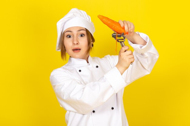 Een vooraanzicht jonge vrouwelijke kok in wit kokkostuum en wit GLB die en oranje wortel op de geel houden schoonmaken