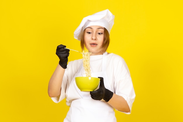 Een vooraanzicht jonge vrouwelijke kok in wit kokkostuum en witte GLB in zwarte handschoenen die groene plaat houden die spaghetti op geel mengen