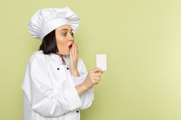 Een vooraanzicht jonge vrouwelijke kok in wit kookkostuum en pet met witte kaart op de groene muur