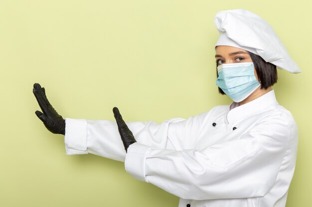 Een vooraanzicht jonge vrouwelijke kok in wit kookpak en pet met handschoenen en een steriel masker met een voorzichtige houding op de groene muur.