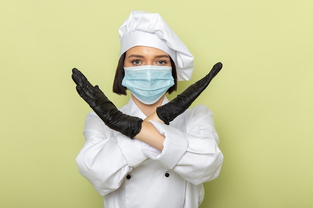 Gratis foto een vooraanzicht jonge vrouwelijke kok in wit kookpak en pet met handschoenen en een steriel masker met verbod pose op de groene muur dame werk voedselkleur