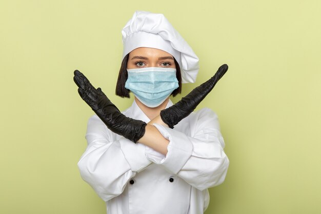 Een vooraanzicht jonge vrouwelijke kok in wit kookpak en pet met handschoenen en een steriel masker met verbod pose op de groene muur dame werk voedselkleur