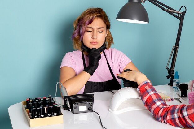 Een vooraanzicht jonge vrouwelijke manicure in roze t-shirt met zwarte handschoenen en zwart masker manicure doen met aarzelende uitdrukking op blauw