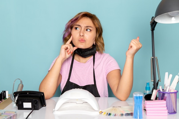 Een vooraanzicht jonge vrouwelijke manicure in roze t-shirt met zwarte handschoenen en zwart masker zit voor de tafel en denkt aan blauw