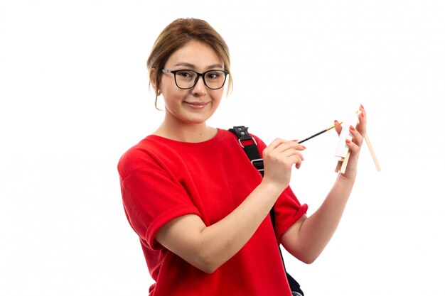 Een vooraanzicht jonge vrouwelijke student die in rode t-shirt zwarte jeans penseel het schilderen houden glimlachend op het wit