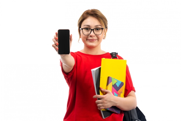 Een vooraanzicht jonge vrouwelijke student die in rode t-shirt zwarte jeans voorbeeldenboeken houdt glimlachend tonend smartphone op het wit