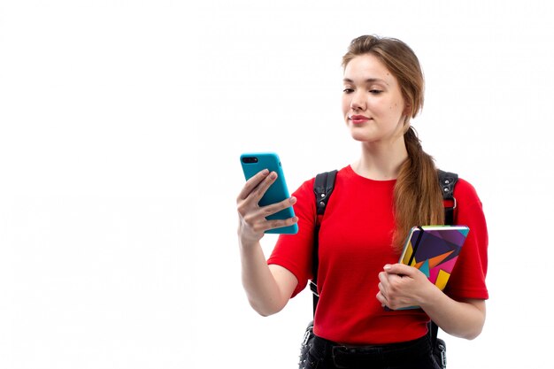 Een vooraanzicht jonge vrouwelijke student in de rode de holdingsvoorbeeldboeken dossiers van de overhemds zwarte zak glimlachend gebruikend haar telefoon op het wit