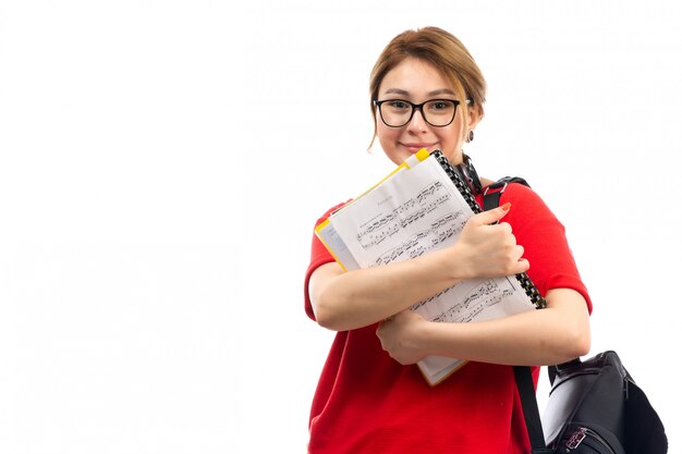 Een vooraanzicht jonge vrouwelijke student in rode t-shirt zwarte jeans die zwarte oortelefoons dragen die notaschrift houden glimlachend op het wit