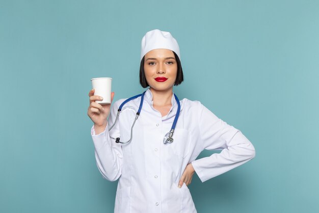 Een vooraanzicht jonge vrouwelijke verpleegster in wit medisch pak en blauwe stethoscoop die plastic glas op de blauwe het ziekenhuisarts van de bureaugeneeskunde houdt