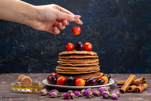 Een vooraanzicht lekkere pannekoeken met kersen binnen witte plaat met bloemen en kaneel op de donkere pannenkoek van het bureaifruit